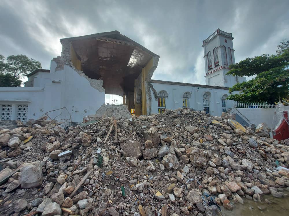 Piles of rubble stand before a severely damaged church. Overhead, dark clouds cast a somber sky.