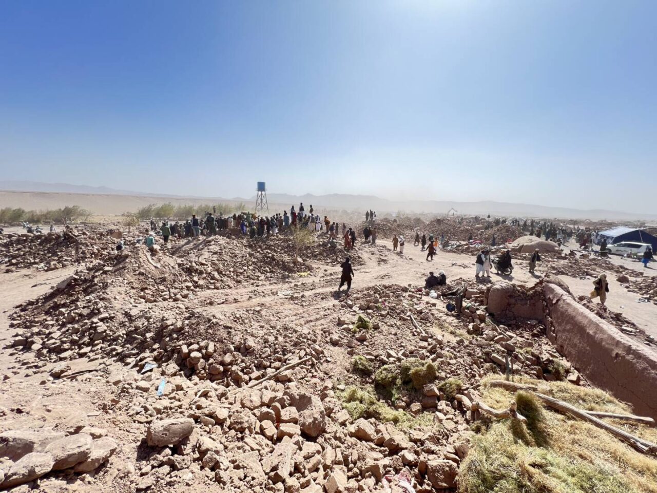 Dust rises from a landscape of rubble and rocks with men standing in the distance.
