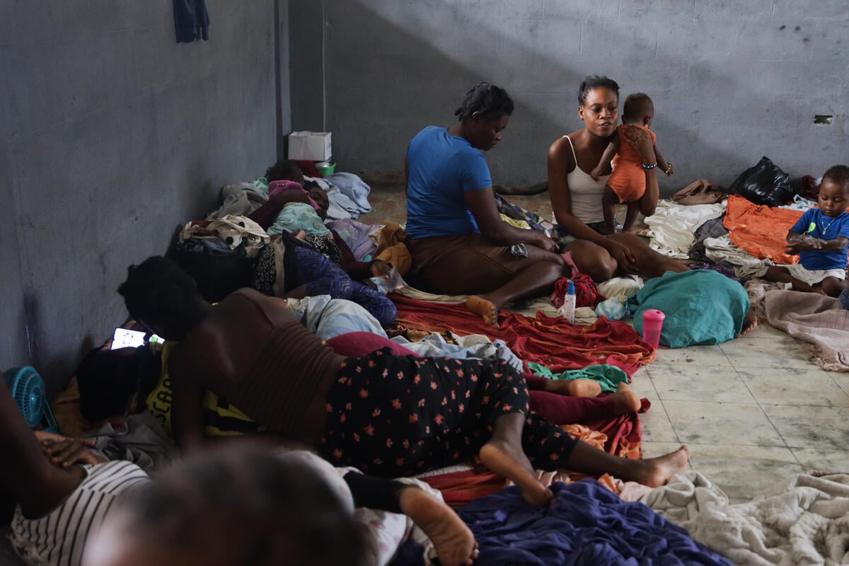 Several people sit and lie on the floor, including one woman holding a baby with a small child nearby.