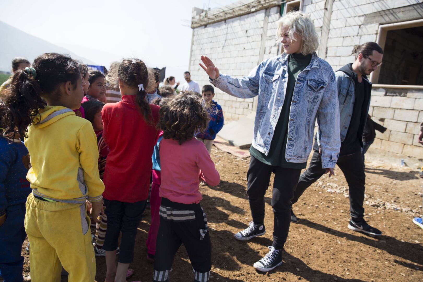 A woman stands outside a brick building and high-fives a crowd of young children. There is a mountain in the background.