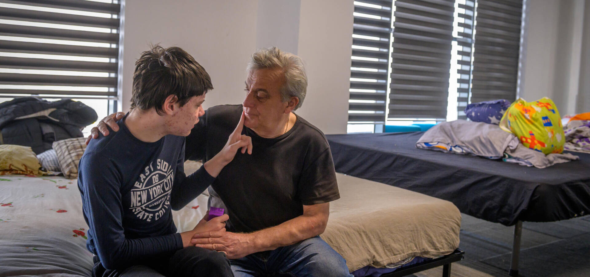 A Ukrainian teenage boy sits next to his father. He places one finger on his father&rsquo;s chin and holds his father&rsquo;s hand with the other. The father's arm is around the boy and he listens intently.