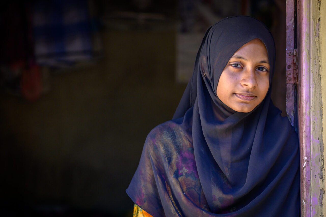 Portrait of a somber young woman wearing a black headscarf standing in a doorway.