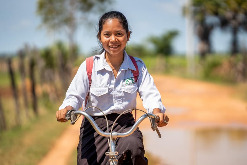 Portrait of a smiling girl on a bike wearing a white shirt, black skirt, and red backpack. There are trees behind her.