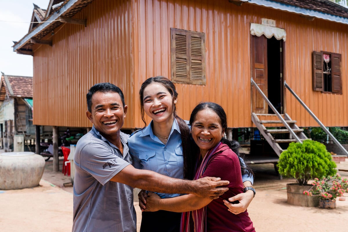 A smiling young woman stands between two adults, most likely her parents, all embracing joyfully in front of a wooden home on stilts. The setting appears to be a rural village.