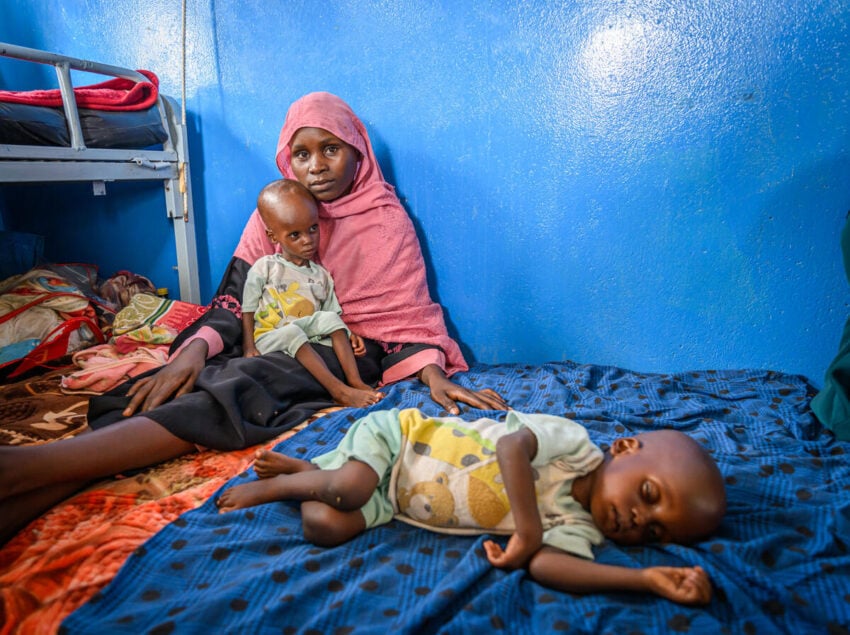 A woman in a pink scarf looks into the camera, holding a malnourished child on her lap while another malnourished child lies beside her.