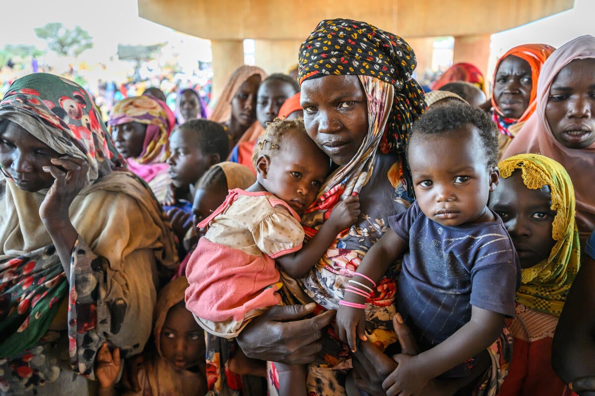 A woman with two young children looks directly into the camera. A crowd of women and children surround her beneath a bridge.