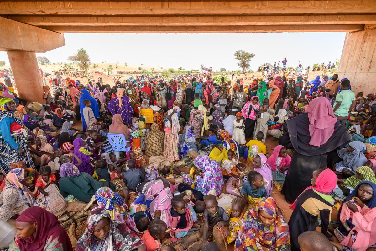 A throng of women and children dressed in colorful garments and headscarves huddle beneath a concrete bridge. The sky is white.