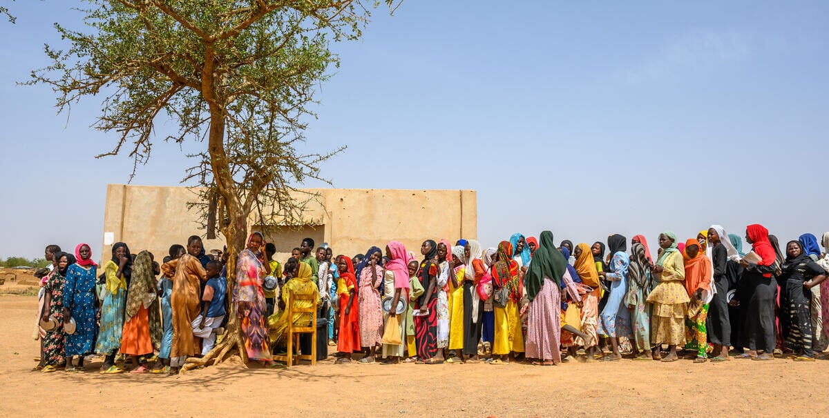 School-aged children stand in line under the sun. A few kids seek shade under a sparse tree.