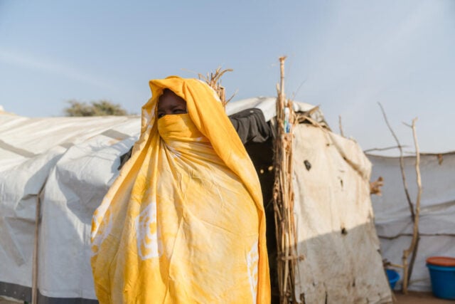 A woman wrapped in a yellow shawl stands in front of makeshift shelters in a displacement camp under a clear sky.