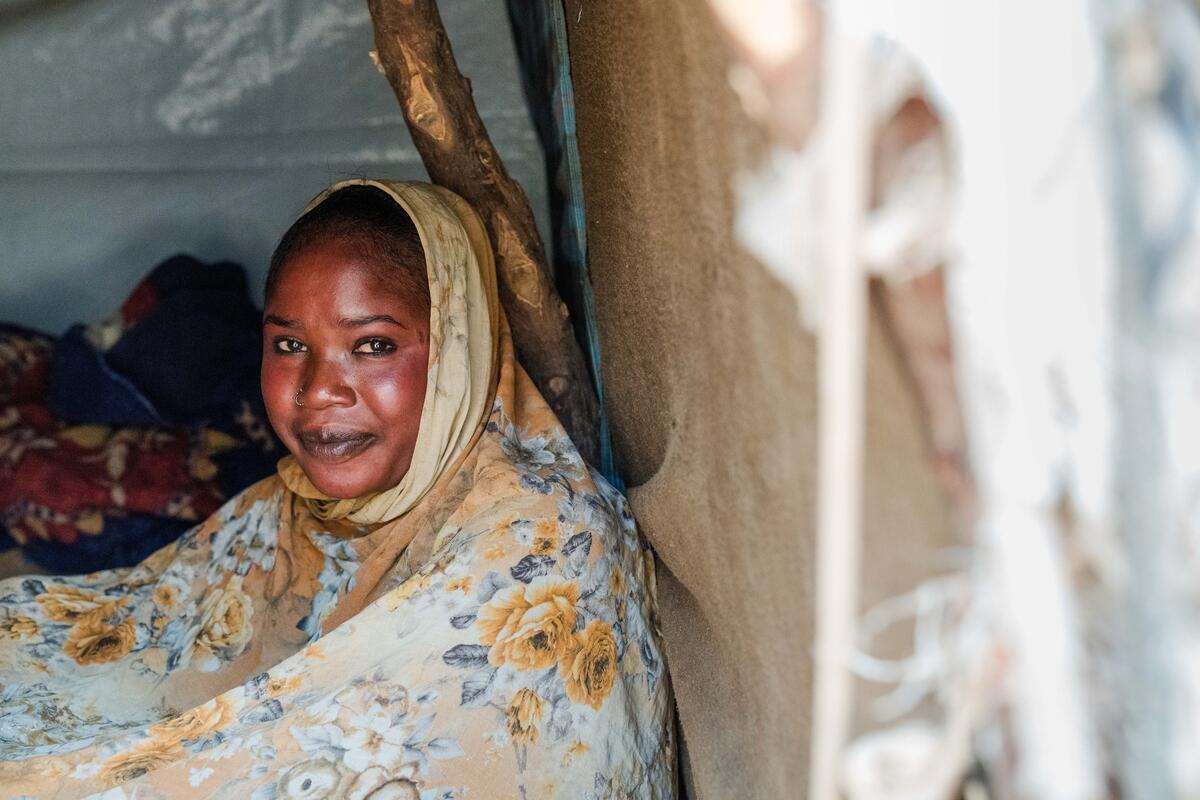 A woman in a floral headscarf sits inside a shelter, looking calmly at the camera.