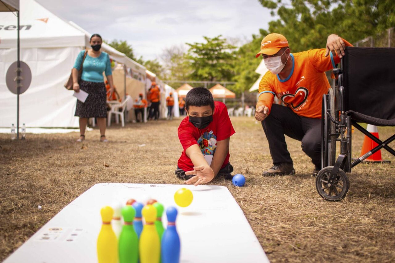 A boy in a red shirt sits on the ground with a World Vision staff person next to him. A woman looks on behind him.