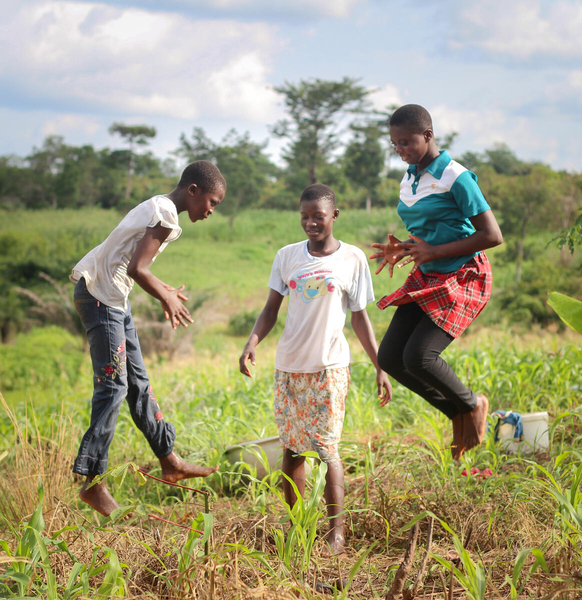 Three Ghanian teenage girls jump in a lush green field while smiling and concentrating on a game.