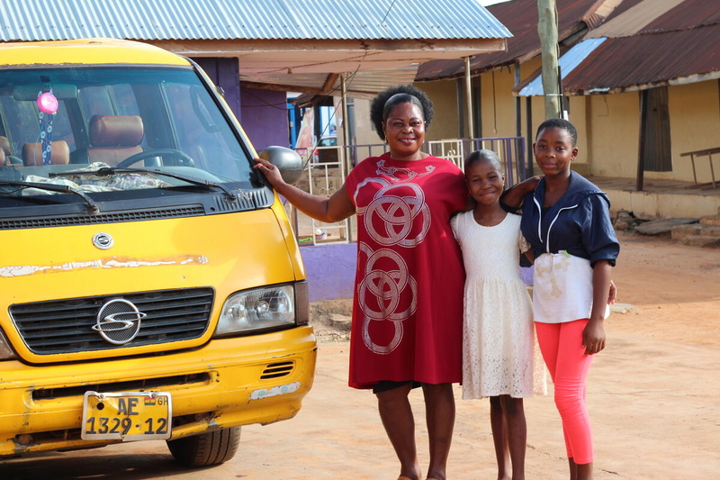 A Ghanian woman in a red dress and two girls stand next to a yellow taxi van smiling.