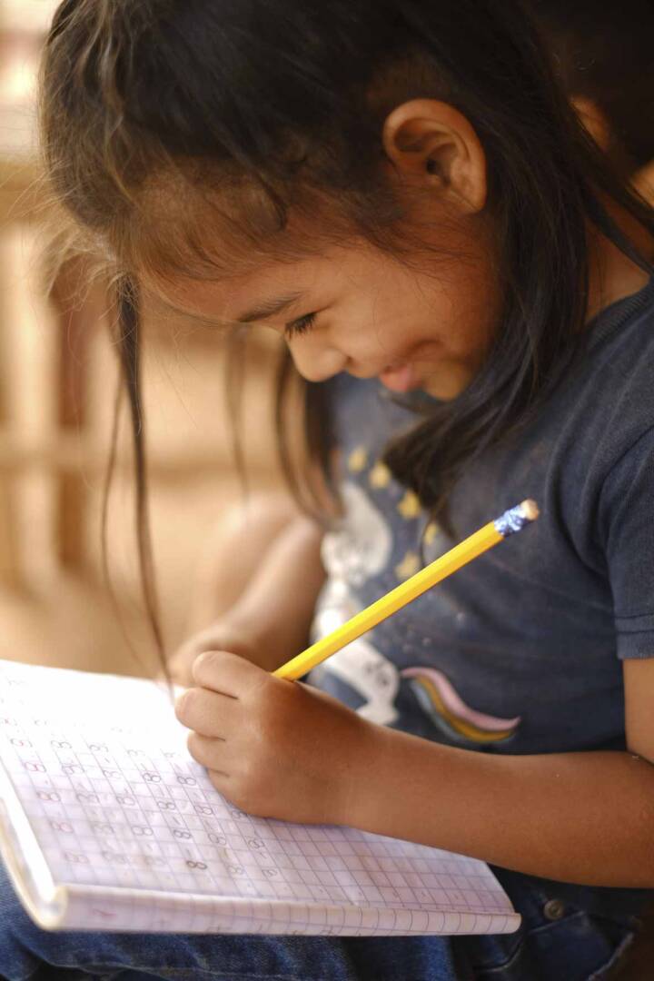 A young Honduran girl sits smiling while holding a yellow pencil and writing in a notebook.