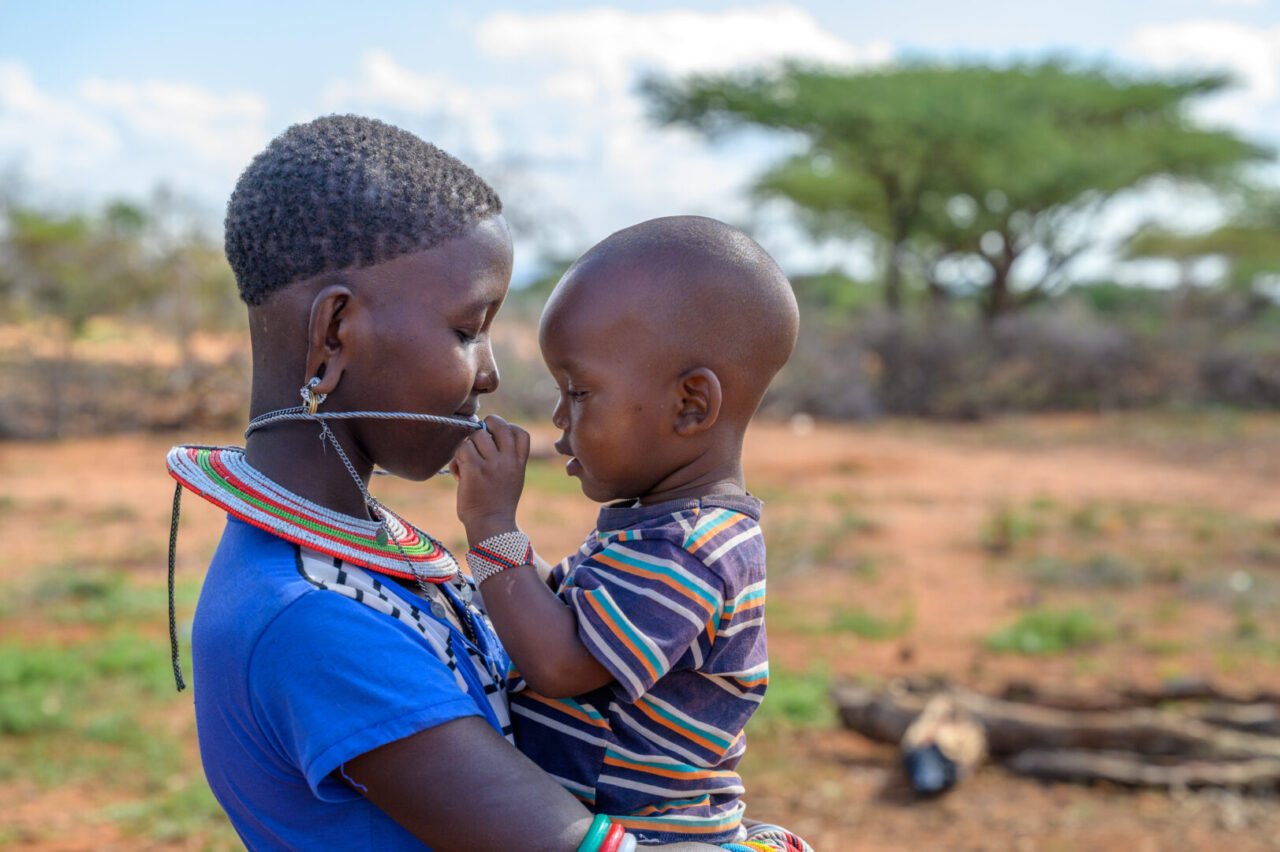 A young woman holds a young boy in her arms. The child plays with the young woman&rsquo;s beaded necklace. 