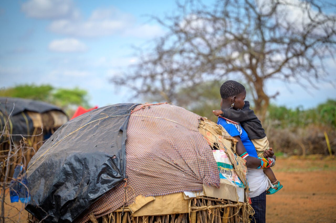 A young woman holds a young boy in her arms as she leans against a makeshift shelter. 