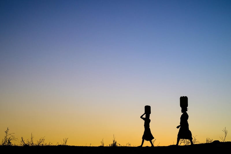 Woman and girl silhouetted against the sunset carry buckets on their heads.