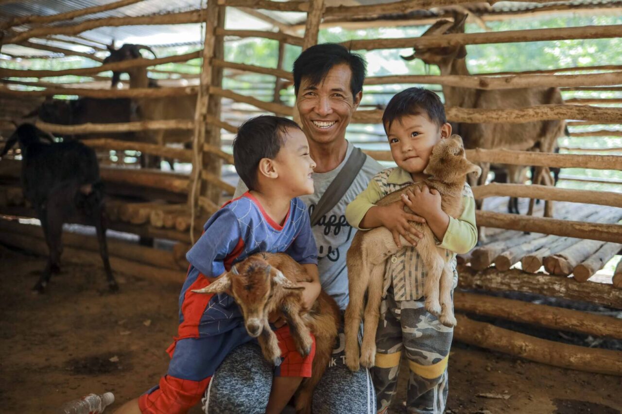 A man smiles standing with two young boys; the children hold goats with other goats in pens behind them.