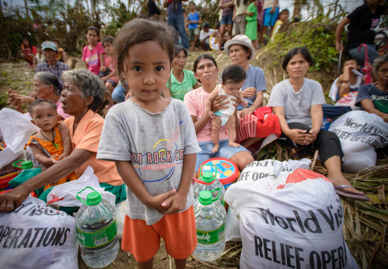 A young girl looks into the camera, with family and other typhoon survivors surrounded by relief supplies.