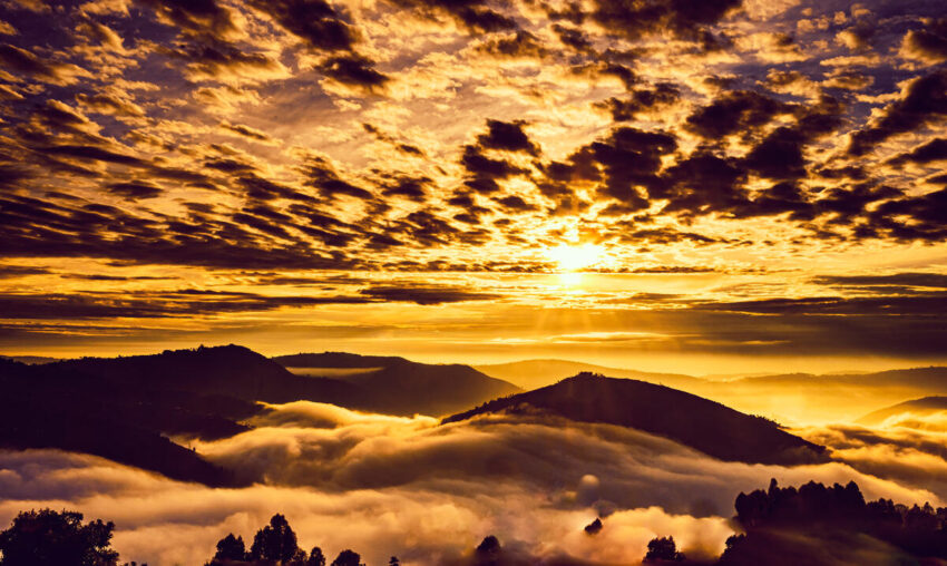 Hilltops emerge through a backdrop of cloudy skies and lowlands, while sunlight filters through the clouds, casting a yellow glow across the land.