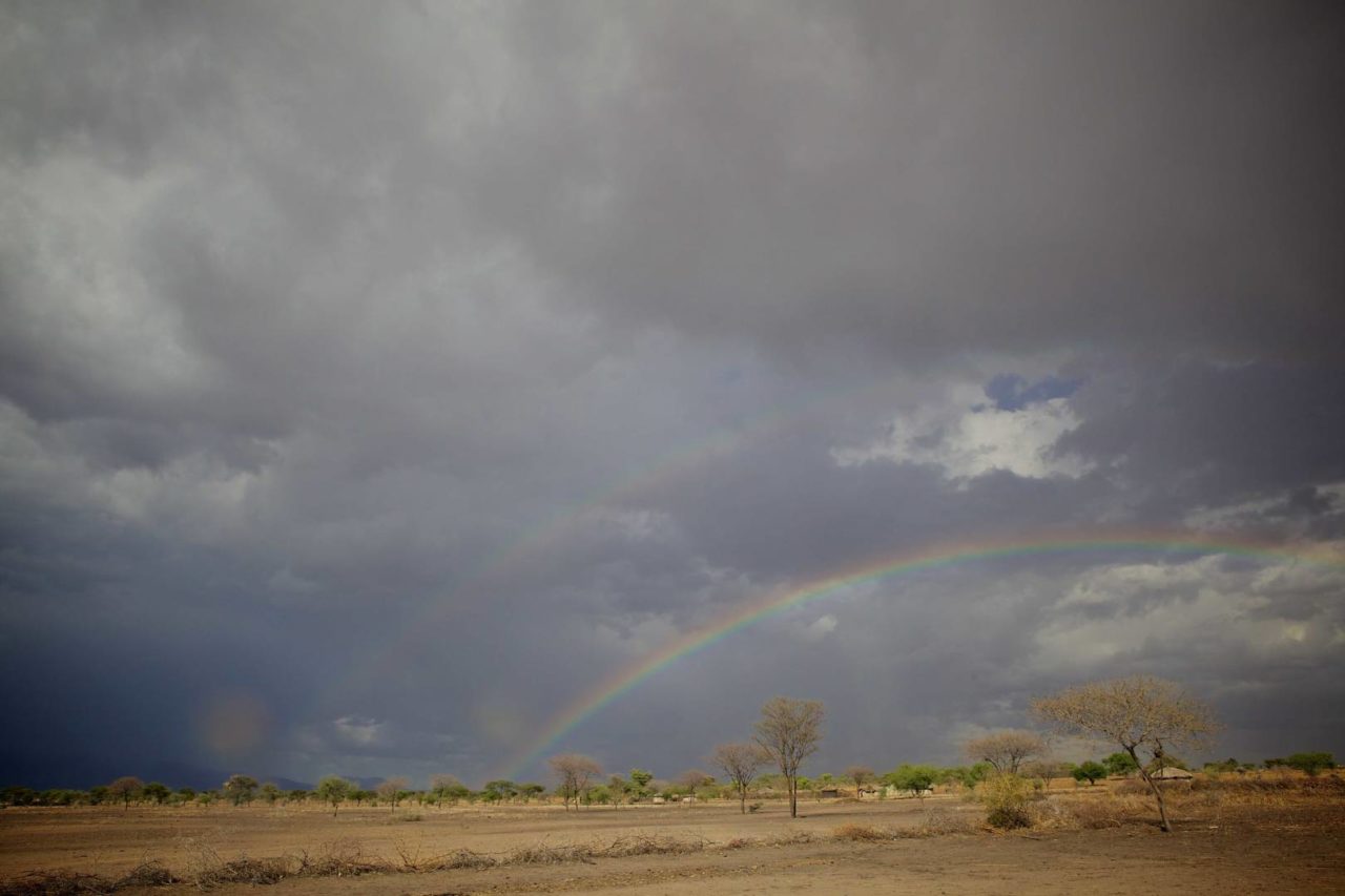 A double rainbow appears through darkened clouds over a rural Tanzanian town.
