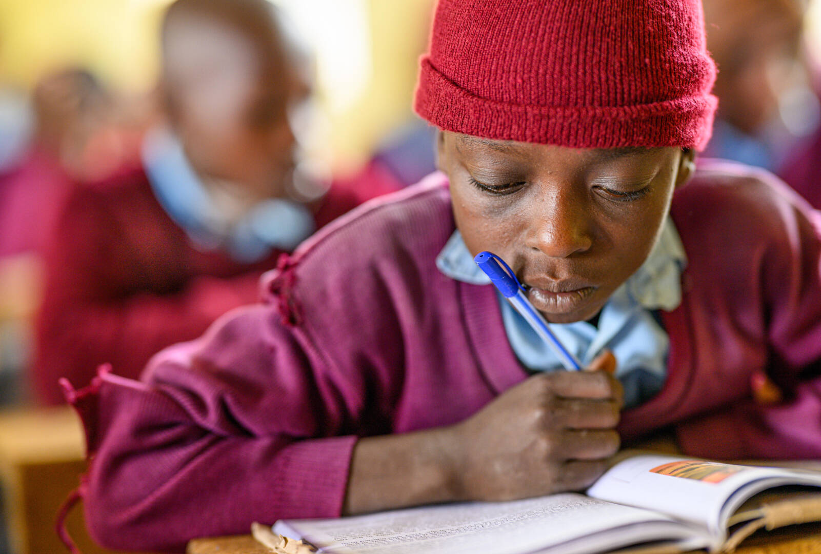 Close-up of a child in a classroom reading a book with his pen leaning against his mouth.