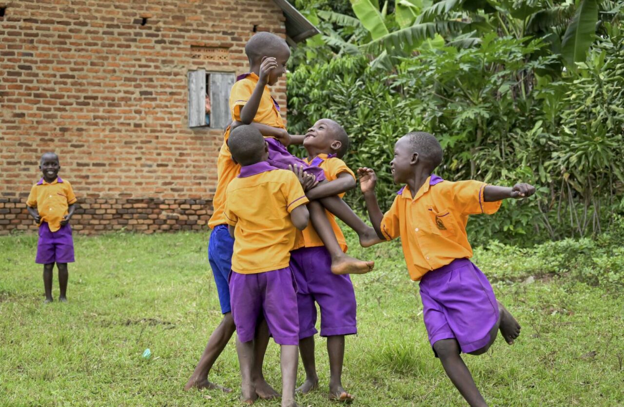 Ugandan boys wearing purple shorts and orange shirts hoist a boy up into the air in triumph as they play soccer in a green yard.