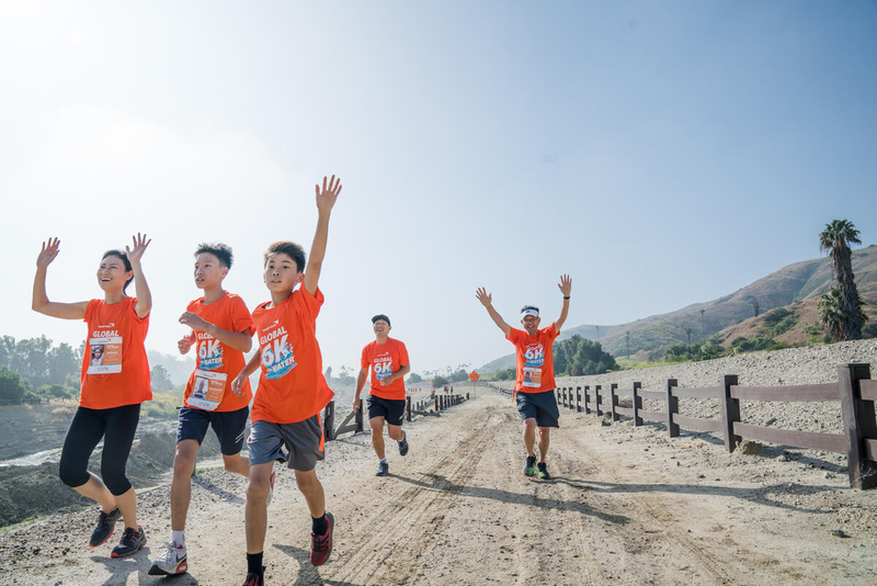 Children and adults run along a dirt trail and wave to a camera. There is a fence to the right, and palm trees and hills beyond it.