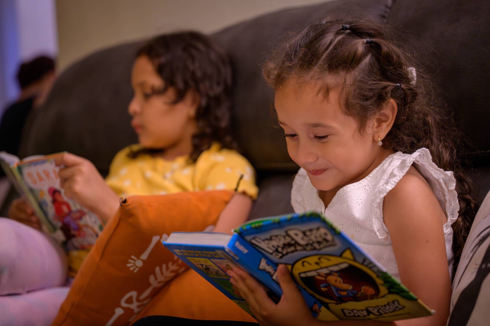 Two young girls read books while sitting on a couch.