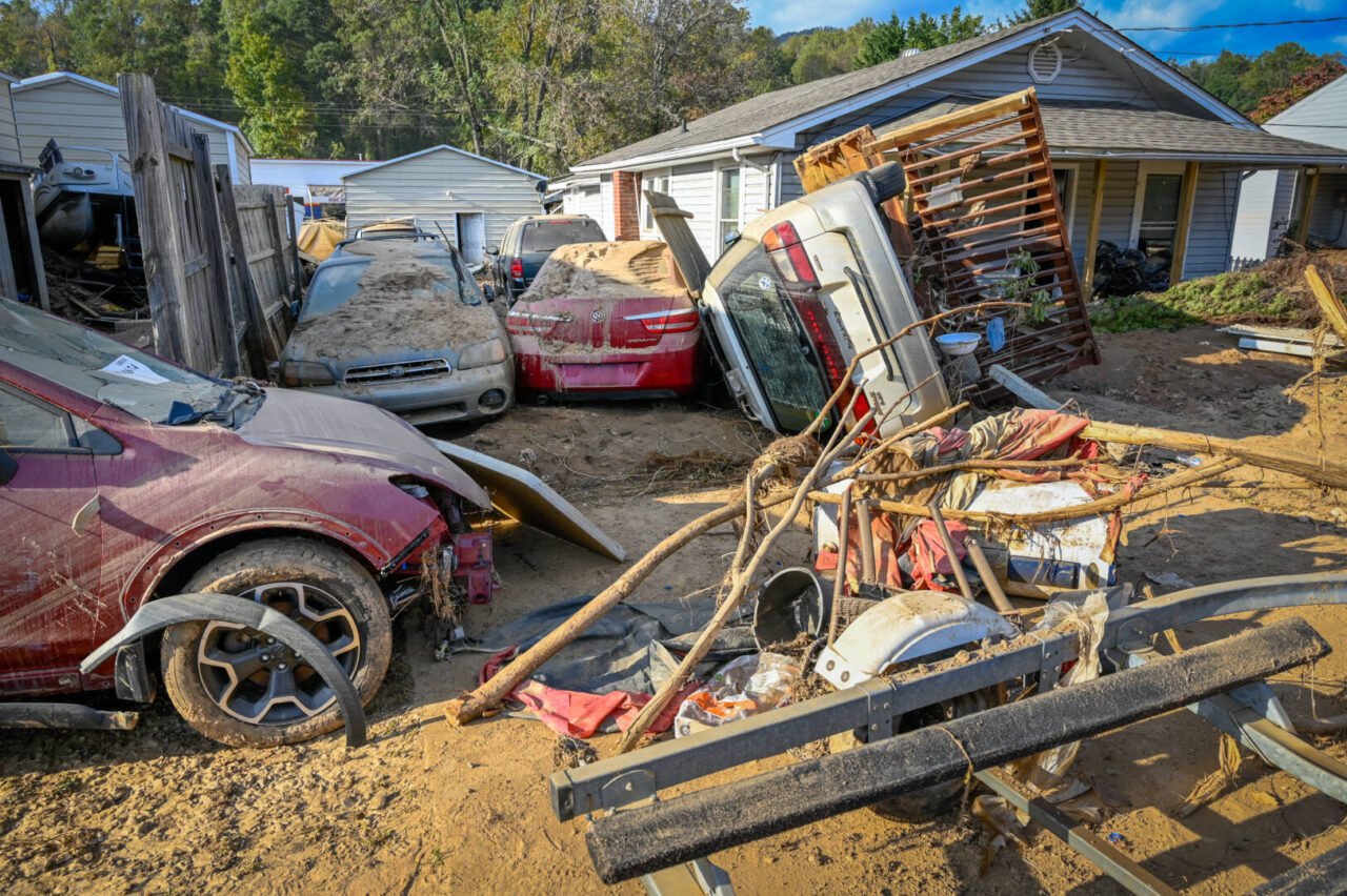 Debris is piled near a damaged doorstep of a home on a sunny day. A car is turned on its side.