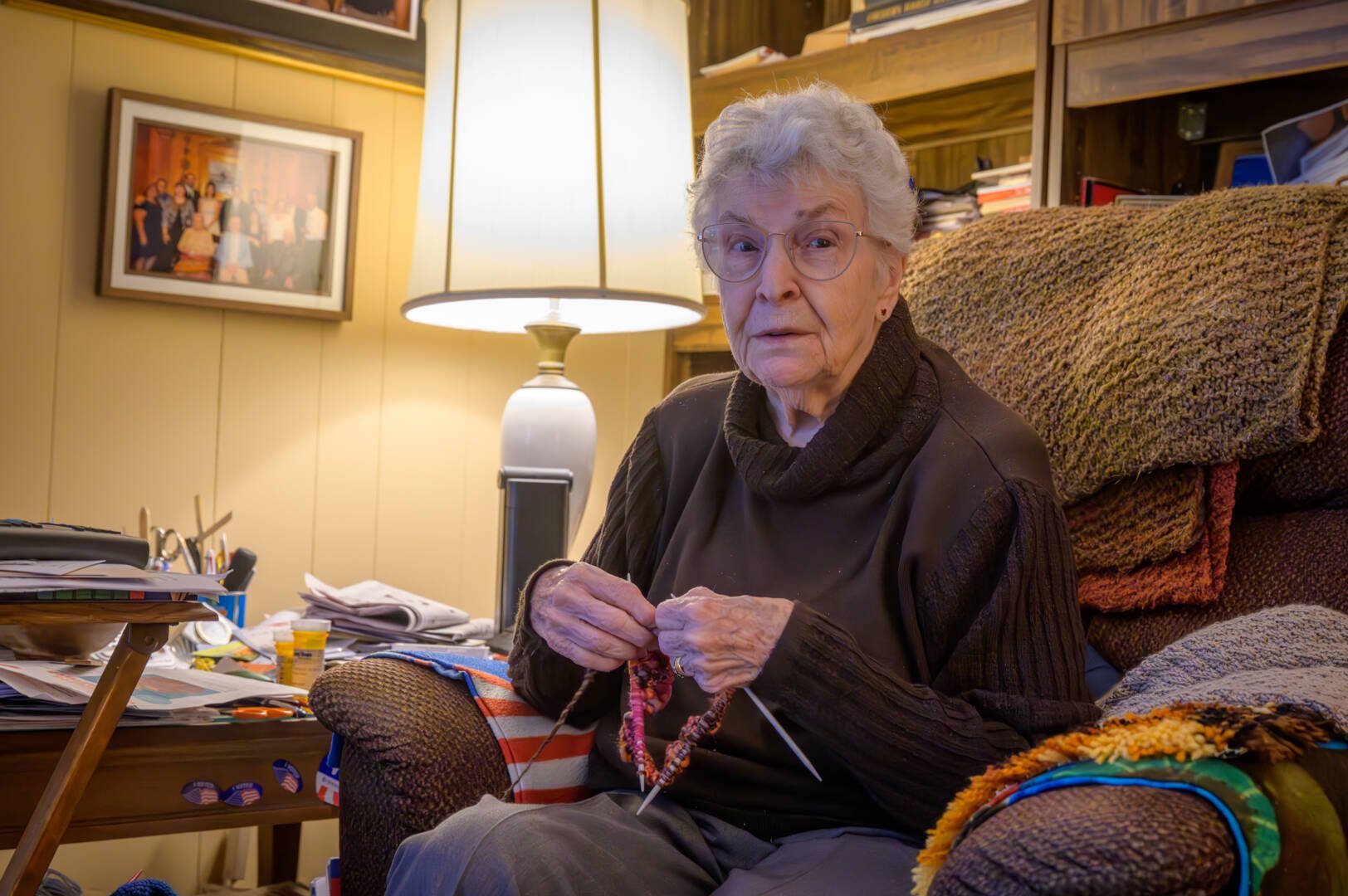 An elderly woman sits in an upholstered chair next to a lamp and knits in the round using four needles.