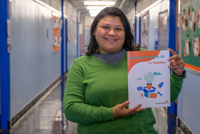 A woman stands in a school hallway holding a photo that reads &ldquo;Classroom Wellbeing Kit for Teachers.&rdquo;