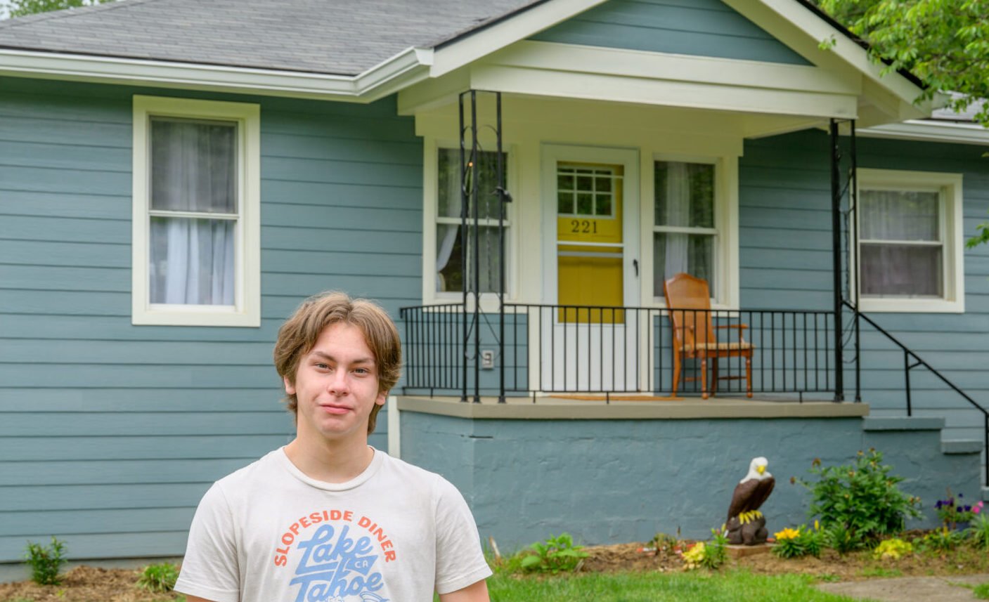 A young man in a white T-shirt stands in front of a blue house with white trim.