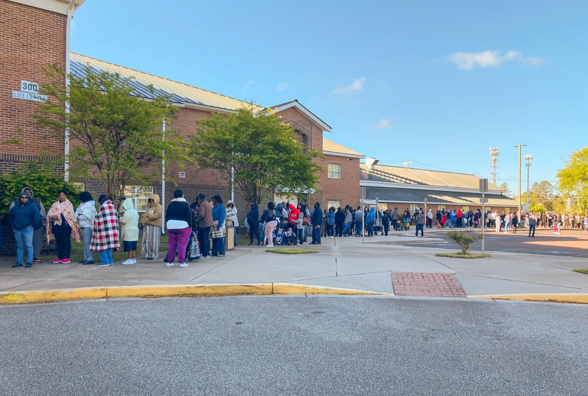 Large group of people stand in a long line outside a brick building on a clear morning, some wrapped in blankets.