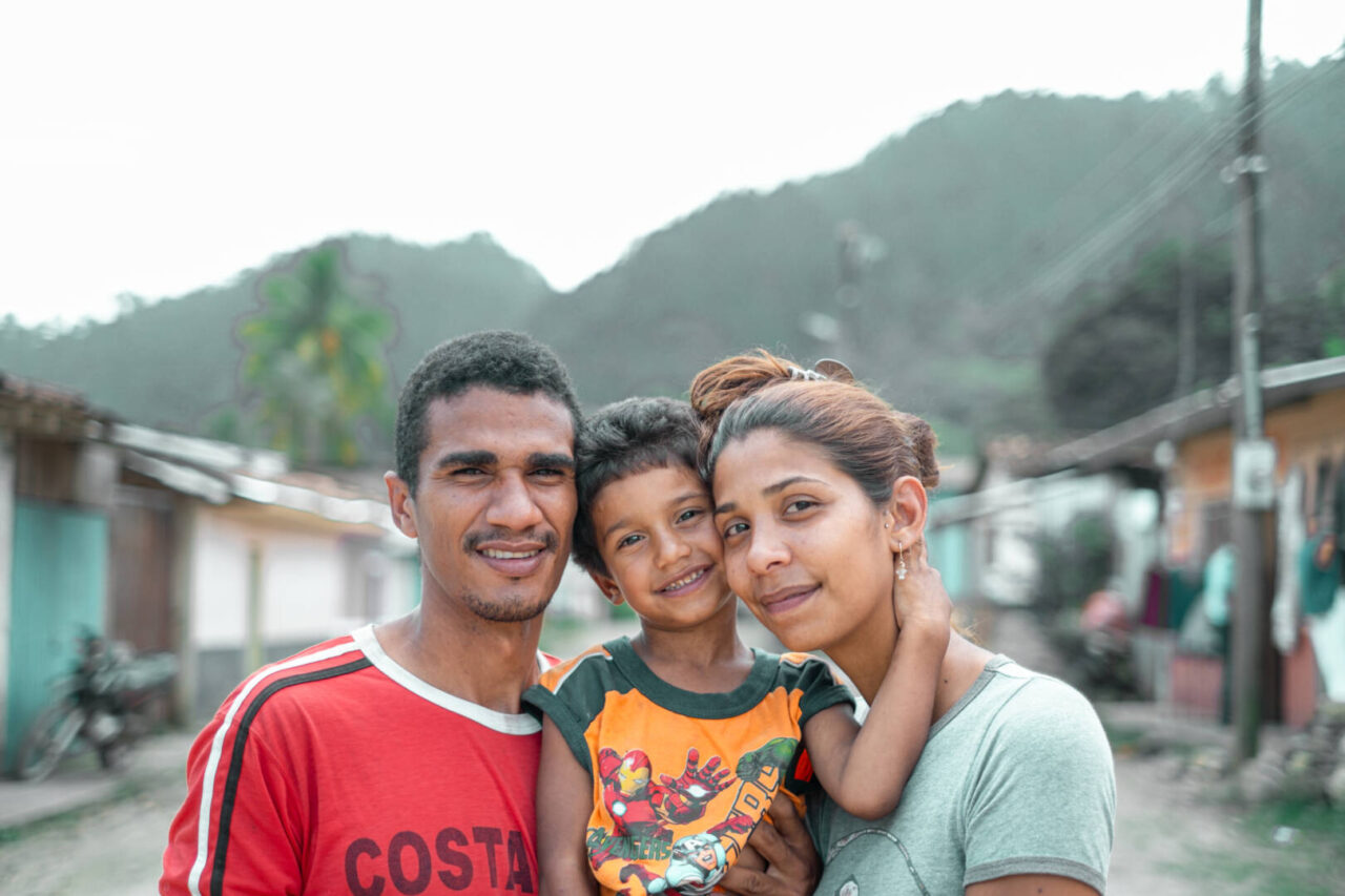 A smiling young boy is nestled between his parents, his arm lovingly wrapped around his mother&rsquo;s neck and face.