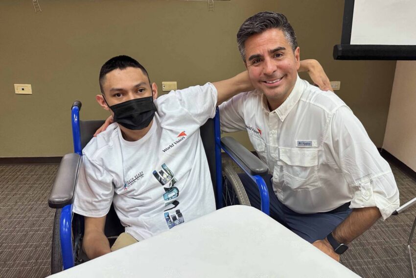 A young man in a white T-shirt sits in a wheelchair with his arm around a middle-aged man in a casual white button-down shirt.