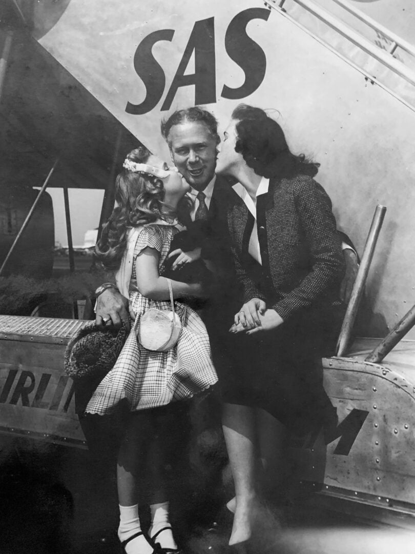 A black-and-white photo of a man in front of an airplane, smiling as a young woman and young girl each kiss him on the cheek.