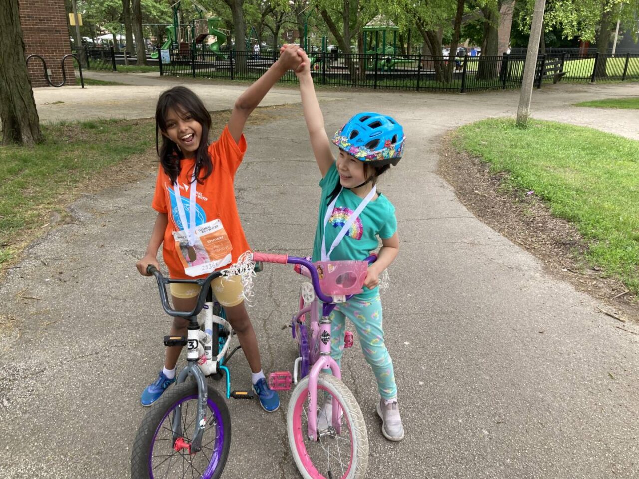 Two little girls on bicycles clasp raised hands over their heads in triumph. They&rsquo;ve done World Vision&rsquo;s Global 6K.