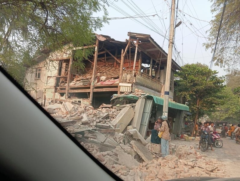 View from a vehicle shows a building collapsed an earthquake, with onlookers standing nearby and some on bikes.