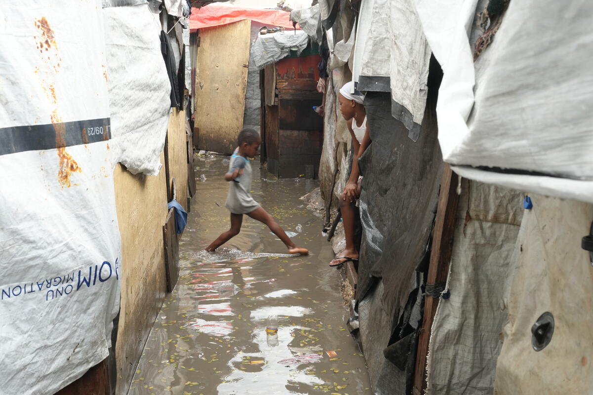 A child skips over a flooded, narrow path in a crowded shelter, while another child looks over their shoulder.