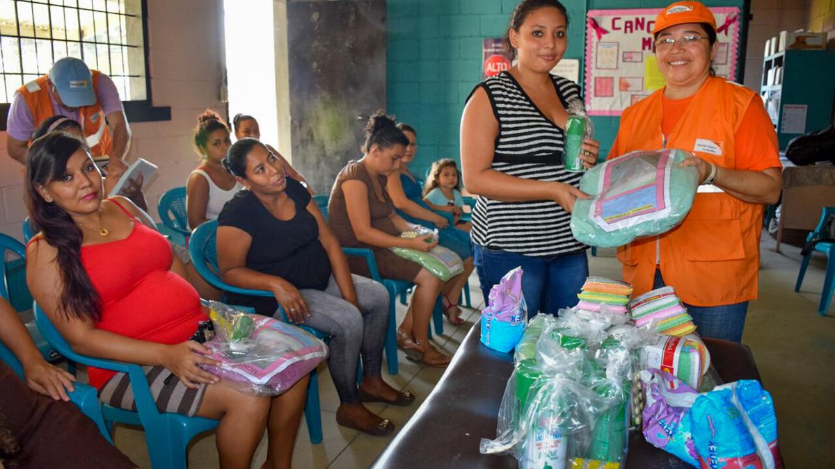 A pregnant woman in a striped shirt and an aid worker holding relief supplies look at the camera. Pregnant women are seated in rows nearby.