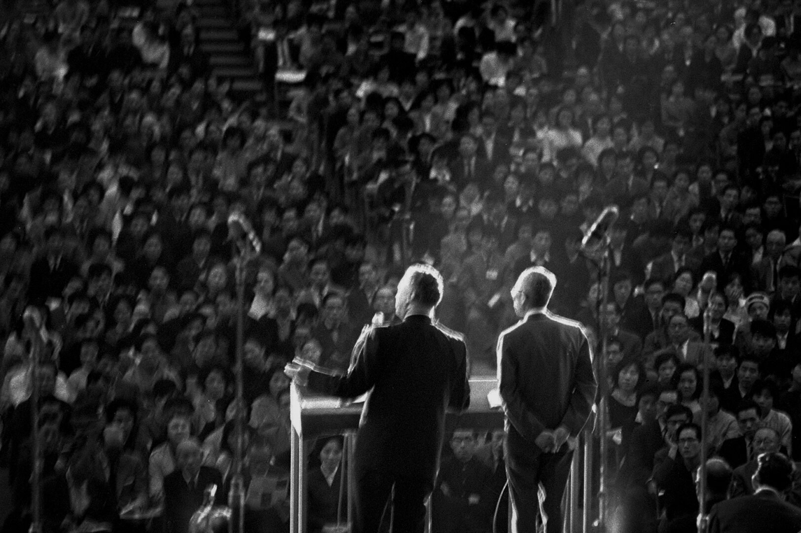 Two men stand on a stage before a large-seated crowd, speaking at a podium under bright lights in a packed auditorium.