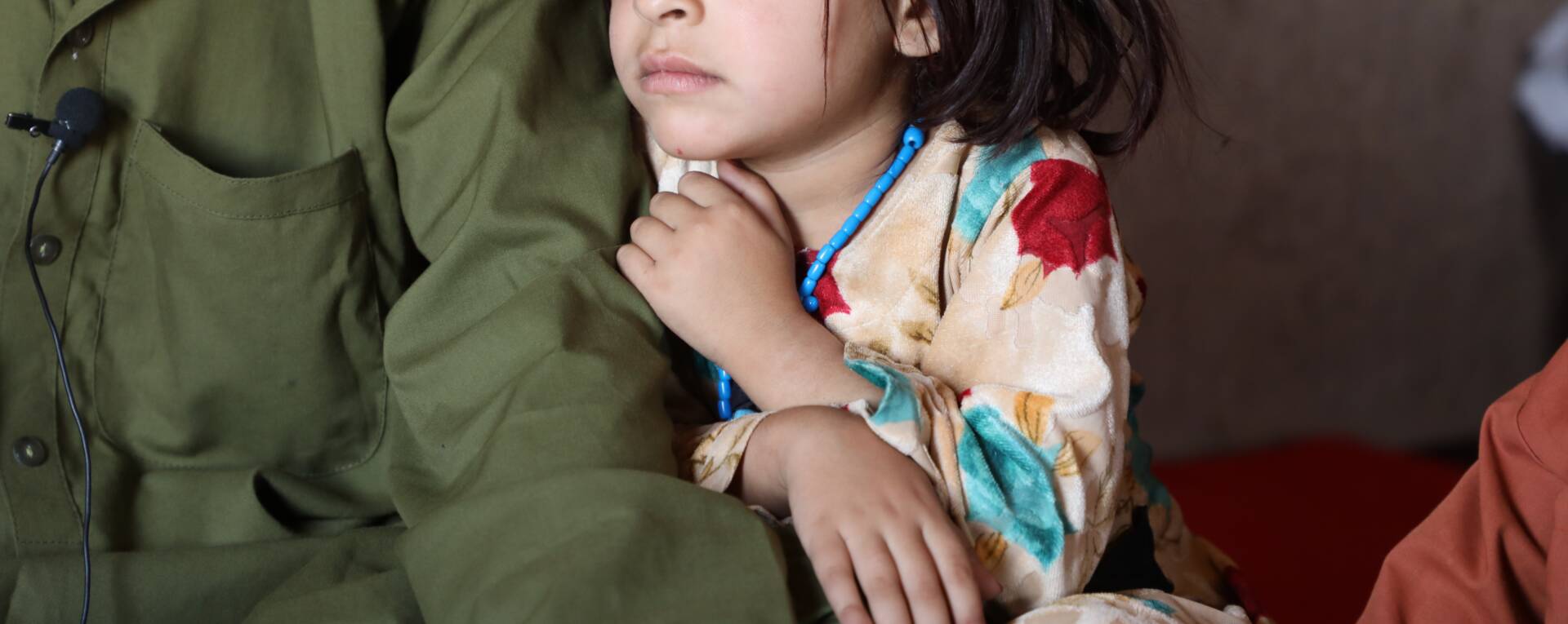 A girl (face partially shown) sits near a man who has a microphone clipped to his olive-green shirt. His face is not shown.