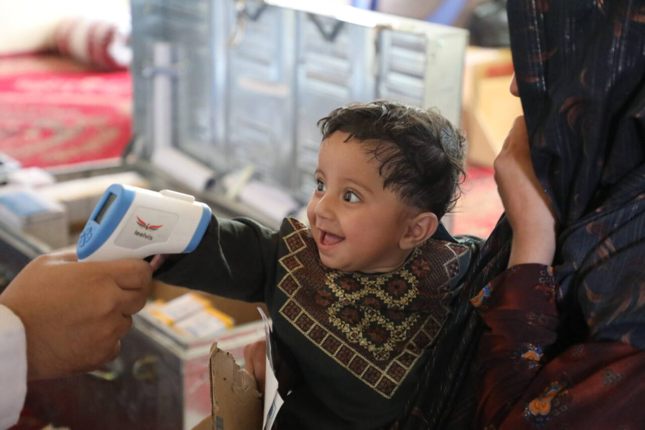 A health worker checks a smiling young Afghan girl&rsquo;s temperature.