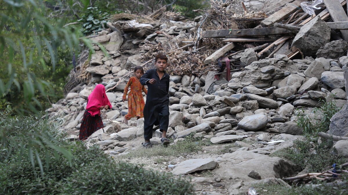 Children walk along a narrow path through the ruins of a destroyed house, with crumbled buildings and scattered rubble.