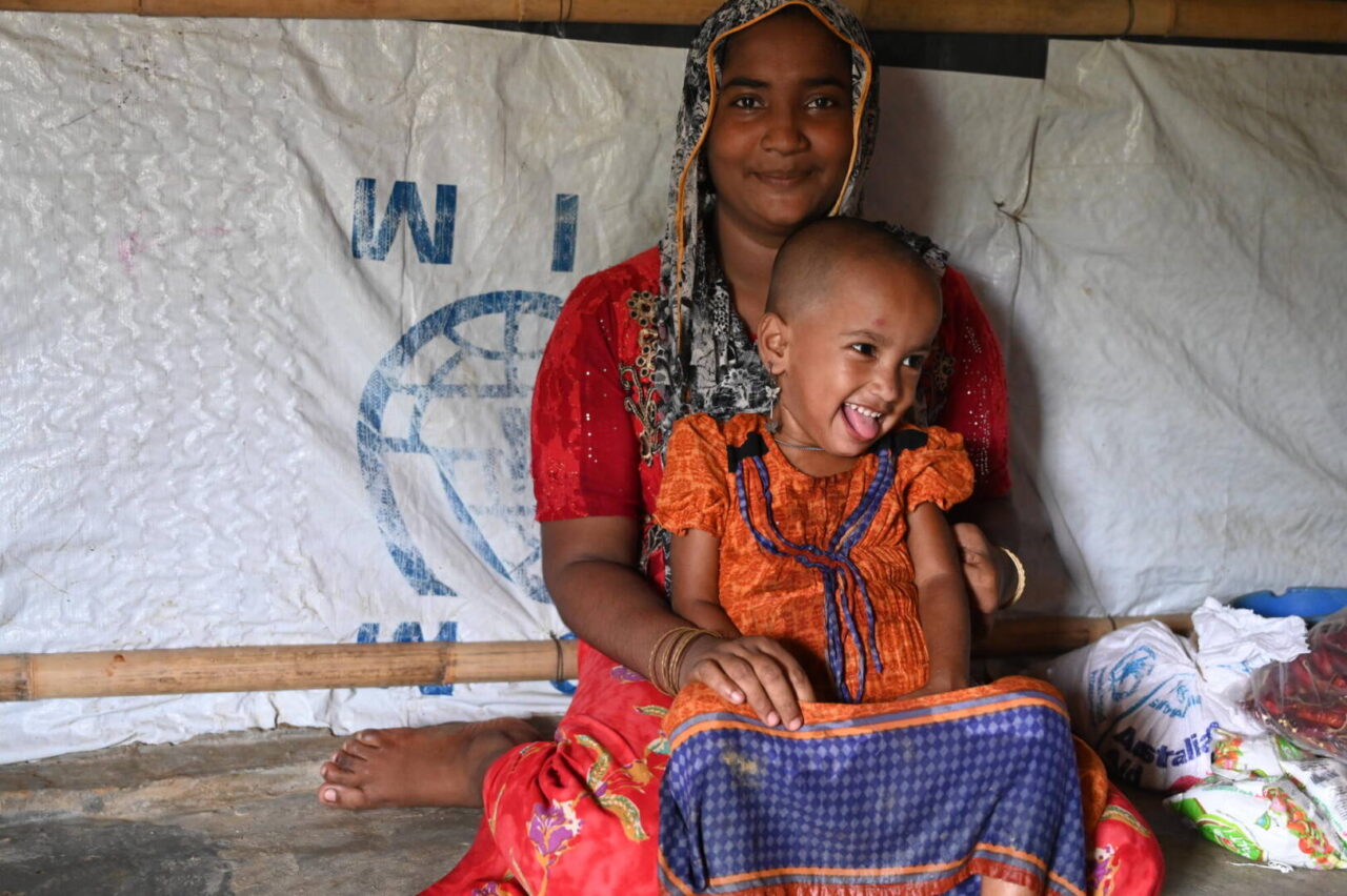 A young Rohingya girl sits on her mother&rsquo;s lap, smiling.