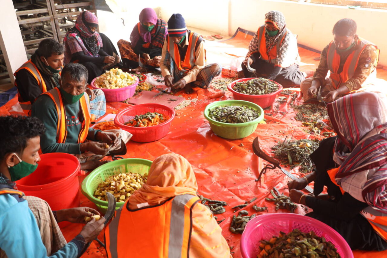 People sit in a circle on the floor around large bins filled with vegetables and other staples during meal preparation for Rohingya refugees. 