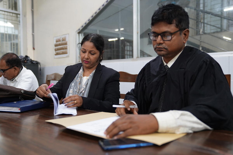 A man in a black robe sits next to a woman in a blazer who is holding a pen. They both look down at documents on the table in front of them.