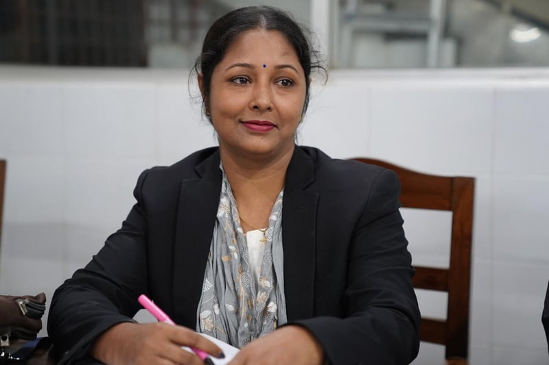 A woman wearing a black blazer with a gray, flowered scarf sits at a table and holds a pen.