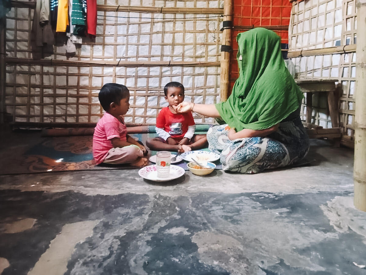A woman in a green scarf sits on the ground feeding a young child, while another child watches them closely.